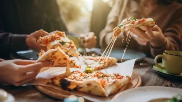 Amigos dividem fatias de pizza puxando o queijo derretido, hábito típico do Brasil, onde o prato costuma ser pedido inteiro e compartilhado à mesa.