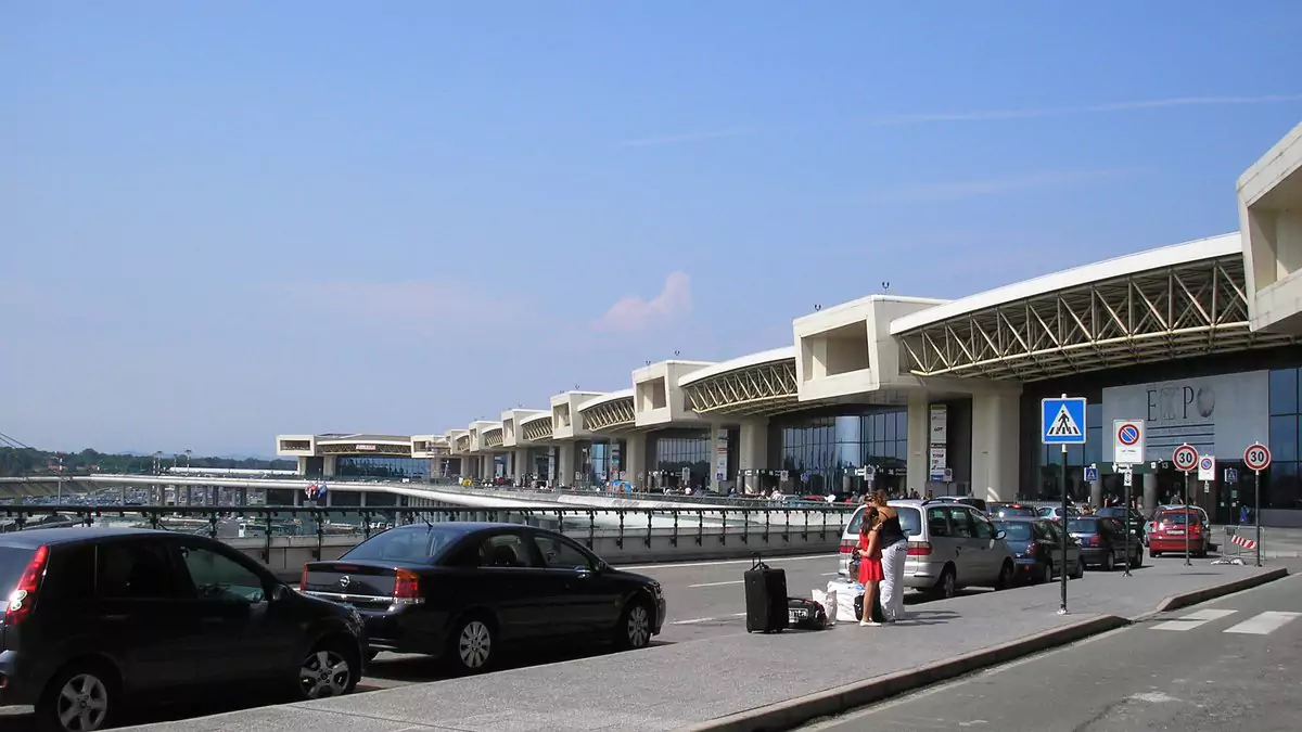 Fachada do Aeroporto de Malpensa, em Milão, com carros e passageiros em frente ao terminal.