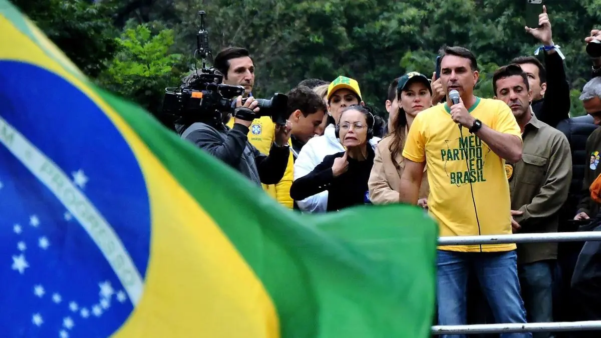 Flávio Bolsonaro discursa diante de apoiadores, com a bandeira do Brasil